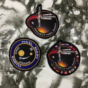 May include: Three circular embroidered patches featuring space-themed designs. Two patches depict a spacecraft with a red trail and the text Project Hail Mary. One patch features a black center with a yellow sun, blue border, and text Hail Mary Earth Grace.