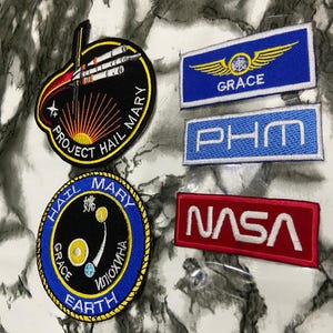 May include: Five embroidered patches including two circular Project Hail Mary mission designs, one blue rectangular patch with yellow wings and the text GRACE, one blue rectangular patch with the text PHM, and one red rectangular NASA logo patch.