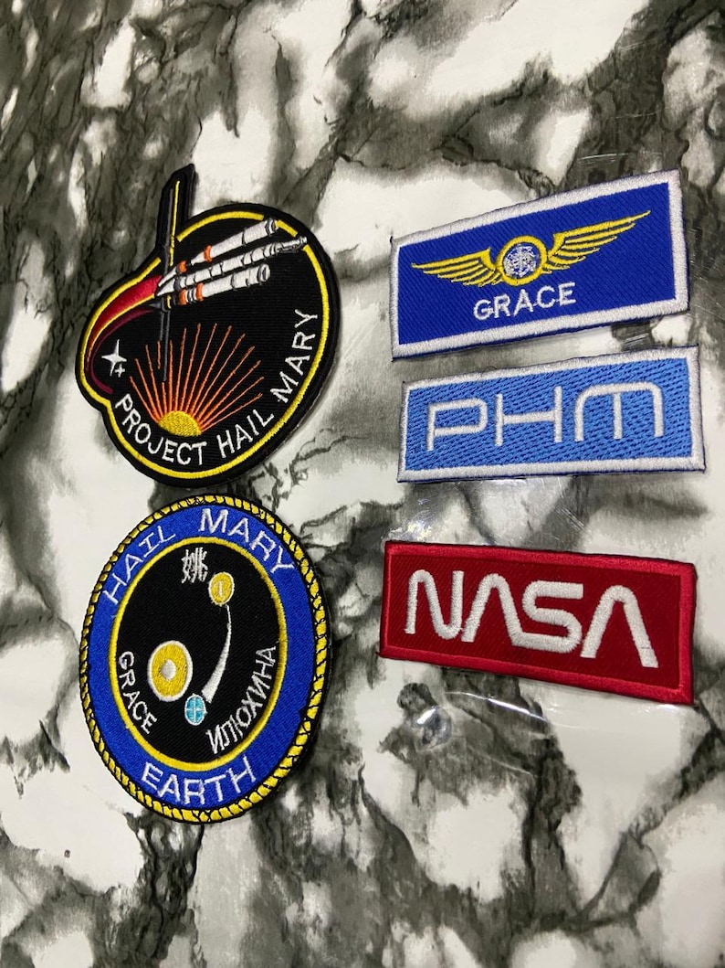 May include: Five embroidered patches including two circular Project Hail Mary mission designs, one blue rectangular patch with yellow wings and the text GRACE, one blue rectangular patch with the text PHM, and one red rectangular NASA logo patch.