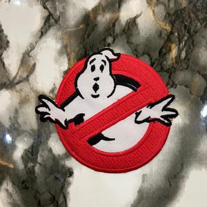 May include: A circular embroidered patch featuring a white ghost character with black outlines and facial features, centered within a red circle that is crossed by a diagonal red bar, all rendered in a classic movie-inspired graphic design.