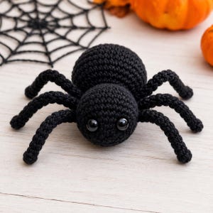 Puede incluir: Una araña de ganchillo negra con cuerpo y cabeza redondos y dos grandes ojos de plástico negro brillante. La araña tiene ocho patas articuladas hechas de hilo negro, que muestran un patrón de punto detallado en toda la estructura.