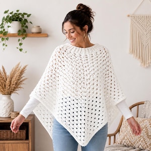 May include: A white crochet poncho featuring an open diamond-shaped stitch pattern and a ribbed knit collar. The garment is worn over a long-sleeved white shirt and blue jeans, creating a layered look with a pointed hemline and textured finish.