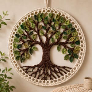 Op de afbeelding: Een ronde wandhanger met een gehaakte boom met een donkerbruine stam en wortels, omringd door talloze kleine groene blaadjes in verschillende tinten, het geheel is ingesloten in een crèmekleurige gehaakte rand als frame.