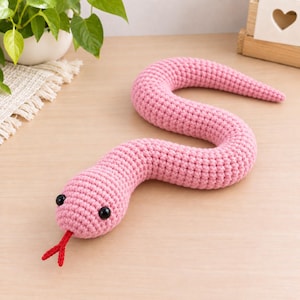 May include: A pink crocheted snake toy featuring a long, winding body made of yarn, two small black bead eyes, and a bright red forked tongue protruding from the front of its rounded head. The texture is uniform and consists of tight stitches.