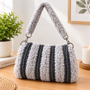 May include: A rectangular crochet handbag featuring alternating vertical stripes of light gray and dark charcoal gray fuzzy yarn. The bag includes a thick, matching textured handle attached with silver-toned metal lobster clasps and rings.