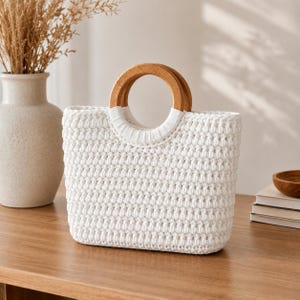 Puede incluir: Un bolso de ganchillo blanco con cuerpo rectangular, patrón de puntos texturizados y dos asas circulares de madera. Las asas están unidas a la parte superior del bolso con conectores de ganchillo blancos, creando una abertura estructurada.