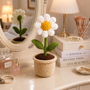 May include: A crocheted daisy flower with white petals and a yellow center, featuring a green stem and two leaves, planted in a textured beige pot with brown soil. The flower is crafted from yarn and stands upright as a decorative item.