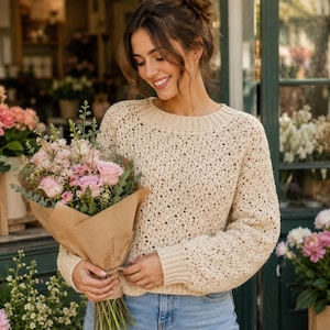 Pode incluir: Um suéter de manga comprida na cor creme com um padrão de malha aberta texturizada, gola redonda canelada e punhos canelados. O suéter é combinado com jeans azuis e segurado junto a um buquê de rosas cor-de-rosa embrulhado em papel pardo.
