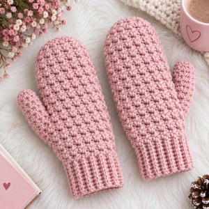 May include: A pair of dusty pink crochet mittens featuring a textured, raised stitch pattern throughout the hand and a ribbed cuff at the wrist. The mittens are crafted with a consistent yarn weight and color, showcasing a uniform, dense construction.