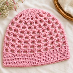 May include: A light pink beanie featuring a prominent open-work grid pattern across the crown and a solid, tightly crocheted band around the base. The headwear is constructed from yarn and displays a consistent, uniform pastel pink color throughout.