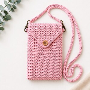 May include: A light pink crochet crossbody bag featuring a rectangular pouch with a triangular flap closure secured by a single brown wooden button. The bag includes a long, matching pink crocheted strap attached with bronze-toned metal clasps.