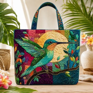 May include: A rectangular quilted tote bag featuring a vibrant, multi-coloured patchwork background. The central design depicts a green hummingbird with orange throat feathers perched on a dark branch against a large, textured yellow moon.