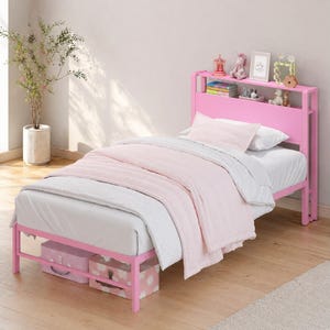 May include: A pink metal twin bed frame with a built-in headboard shelf featuring various small decorative items. The bed is dressed with a white mattress, a light pink blanket, and a white pillow. Two patterned storage boxes sit underneath.