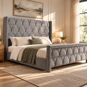 May include: A gray upholstered bed frame featuring a tall, wingback headboard and a low-profile footboard, both decorated with a scalloped, tufted pattern and silver nailhead trim. The bed is dressed with white pillows and a green throw blanket.