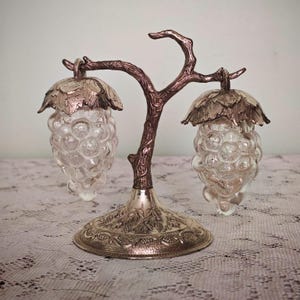 Puede incluir: Un soporte de metal con forma de rama de árbol y base texturizada, que sostiene dos saleros y pimenteros de vidrio colgantes. Cada recipiente tiene forma de racimo de uvas con una tapa metálica decorada con un patrón de hojas en la parte superior.