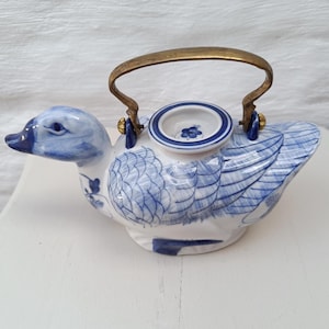 Può includere: Una teiera in ceramica bianca a forma di anatra con dettagli di piume dipinti a mano in blu e un motivo floreale blu sul coperchio. La teiera presenta un manico curvo in ottone fissato sulla parte superiore, con becco e occhi blu.