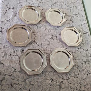Op de afbeelding: Een set van zes zilverkleurige metalen borden, elk met een uitgesproken achthoekige vorm met een glad, reflecterend oppervlak en een opstaande rand. De borden zijn in een cirkelvormig patroon gerangschikt en vertonen een uniforme metalen afwerking.