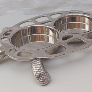 May include: A silver-toned metal serving dish shaped like a turtle, featuring two circular, smooth-bottomed bowls integrated into the shell. The turtle's head and four legs are textured with a scale-like pattern, while the shell has open cutouts.