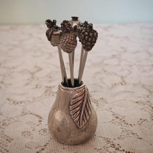 Può includere: Un set di quattro stuzzicadenti da cocktail in metallo argentato con terminali testurizzati a forma di frutta, tra cui un ananas, una ghianda e altre forme botaniche, inseriti in un supporto metallico a forma di pera con una foglia in rilievo.