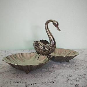 Pode incluir: Um prato de servir em metal prateado com uma figura central de cisne com pescoço arqueado. O cisne é ladeado por duas taças rasas em forma de concha com bordas recortadas e pés pequenos, criando uma peça de servir unificada e composta.
