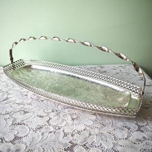 Può includere: Un vassoio da portata rettangolare color argento caratterizzato da un bordo con motivo a reticolo intricato e un design floreale centrale in rilievo. Il vassoio include un'unica maniglia in metallo ritorto e rialzata fissata alle estremità.