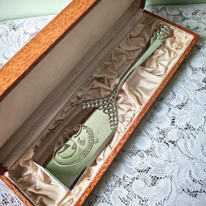 Puede incluir: Un servidor de pescado de tono plateado con acabado pulido, que presenta un diseño de pez grabado en la hoja y un patrón texturizado similar a escamas en el mango, presentado dentro de una caja rectangular de madera forrada con tela color crema.