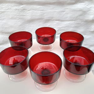 May include: A set of five matching dessert bowls featuring deep red glass basins supported by clear glass pedestal bases. Each bowl has a wide, circular opening and a smooth, rounded silhouette, arranged in a cluster on a flat surface.