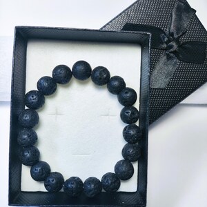 Pode incluir: Uma pulseira circular composta por contas redondas e porosas de pedra de lava preta. A pulseira é apresentada dentro de uma caixa de presente retangular preta com superfície texturizada e um laço de fita preta na tampa, sobre um suporte branco.