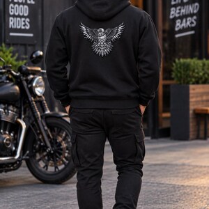 K&ouml;nnte beinhalten: Ein schwarzer Kapuzenpullover mit einer detaillierten wei&szlig;en Grafik eines Adlers mit ausgebreiteten Fl&uuml;geln auf dem R&uuml;cken, kombiniert mit einer schwarzen Cargohose und wei&szlig;en Turnschuhen. Der Hoodie hat lange &Auml;rmel und gerippte B&uuml;ndchen.