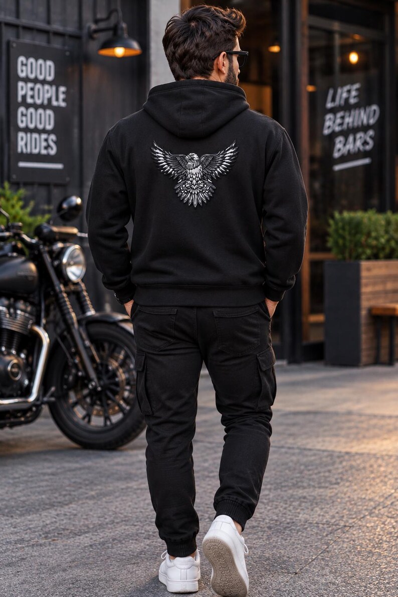 K&ouml;nnte beinhalten: Ein schwarzer Kapuzenpullover mit einer detaillierten wei&szlig;en Grafik eines Adlers mit ausgebreiteten Fl&uuml;geln auf dem R&uuml;cken, kombiniert mit einer schwarzen Cargohose und wei&szlig;en Turnschuhen. Der Hoodie hat lange &Auml;rmel und gerippte B&uuml;ndchen.