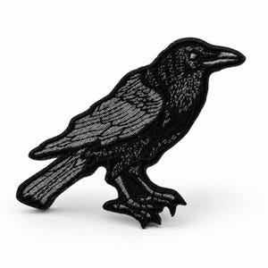 May include: An embroidered iron-on patch featuring a raven in profile. The bird is rendered in shades of black and dark gray thread, showcasing detailed stitching on the wings, feathers, and beak. The design has a solid black border outline.