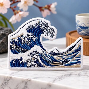 May include: A rectangular embroidered patch featuring a detailed depiction of a large, curling ocean wave in shades of deep blue, white, and tan. The design includes intricate stitching that captures the movement of water and a mountain peak.