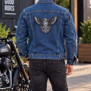 K&ouml;nnte beinhalten: Eine blaue Jeansjacke mit einem gro&szlig;en gestickten Aufn&auml;her eines Adlers mit ausgebreiteten Fl&uuml;geln auf dem R&uuml;cken. Die Jacke hat einen klassischen Kragen, lange &Auml;rmel mit gekn&ouml;pften Manschetten und zwei Brusttaschen. Das Motiv ist schwarz-wei&szlig;-grau.