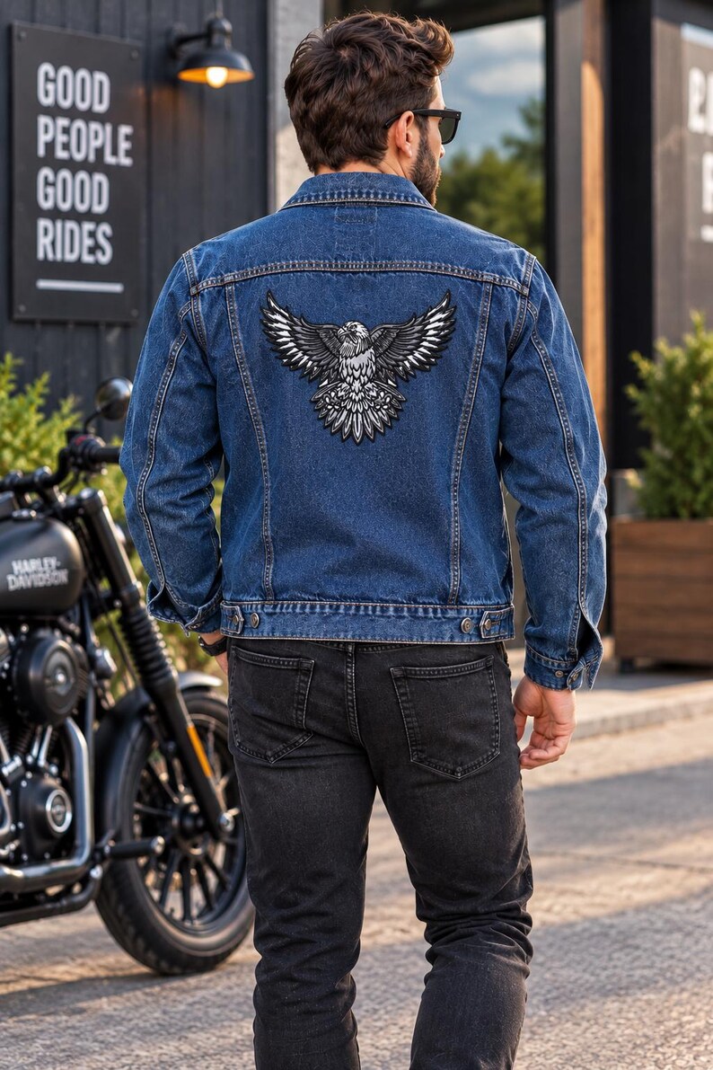 K&ouml;nnte beinhalten: Eine blaue Jeansjacke mit einem gro&szlig;en gestickten Aufn&auml;her eines Adlers mit ausgebreiteten Fl&uuml;geln auf dem R&uuml;cken. Die Jacke hat einen klassischen Kragen, lange &Auml;rmel mit gekn&ouml;pften Manschetten und zwei Brusttaschen. Das Motiv ist schwarz-wei&szlig;-grau.
