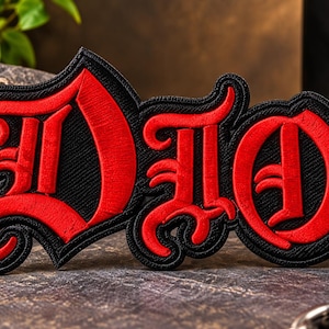 May include: A black and red embroidered patch featuring the text DIO in a stylized gothic font. The letters are rendered in a vibrant red thread with a thick black border, creating a bold contrast. The patch has a textured surface and edges.