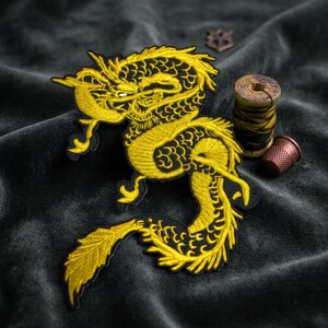 May include: An embroidered patch featuring a golden dragon with intricate scale details and flowing whiskers. The dragon is depicted in a curved, serpentine pose with a long, feathered tail. Beside the patch sits a wooden spool of thread and a thimble.