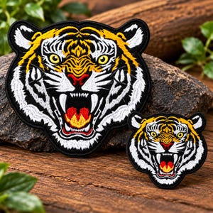 May include: Two embroidered tiger head patches featuring a snarling expression with bared teeth. The design incorporates vibrant yellow, black, white, and red thread colors, showcasing detailed fur patterns and a fierce, open-mouthed tiger face.