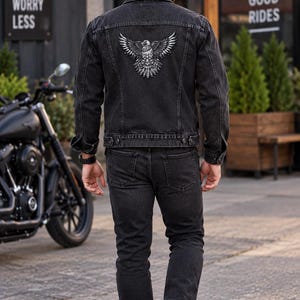 K&ouml;nnte beinhalten: Eine schwarze Jeansjacke mit einer detaillierten wei&szlig;en Adlergrafik auf dem R&uuml;cken, kombiniert mit einer passenden schwarzen Jeans und wei&szlig;en Turnschuhen. Die Jacke hat einen klassischen Kragen, gekn&ouml;pfte Manschetten und eine Knopfleiste.