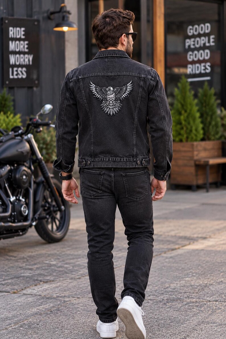 K&ouml;nnte beinhalten: Eine schwarze Jeansjacke mit einer detaillierten wei&szlig;en Adlergrafik auf dem R&uuml;cken, kombiniert mit einer passenden schwarzen Jeans und wei&szlig;en Turnschuhen. Die Jacke hat einen klassischen Kragen, gekn&ouml;pfte Manschetten und eine Knopfleiste.