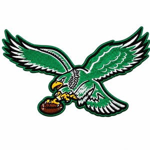 May include: An embroidered patch featuring a green eagle with white and black wing details, a yellow beak, and yellow talons clutching a brown American football. The design is outlined in a thick black border against a plain white background.