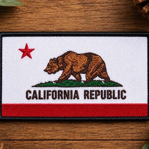 May include: A rectangular embroidered patch featuring the California state flag design. It displays a brown grizzly bear walking on a green patch of grass, a red five-pointed star, and the text CALIFORNIA REPUBLIC in black capital letters.