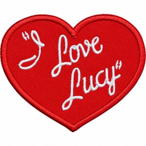 May include: A heart-shaped embroidered patch in bright red with a stitched border. The text "I Love Lucy" is embroidered in white cursive script across the center of the heart, featuring quotation marks at the beginning and the end of the phrase.