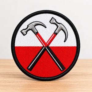 May include: A circular embroidered patch featuring a white upper half and a red lower half. Two crossed hammers with silver heads and red and black handles are centered on the patch. The entire design is bordered by a thick black stitched edge.