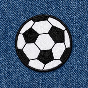 May include: A circular embroidered patch featuring a classic black and white soccer ball pattern with a traditional pentagon and hexagon design. The patch has a thick black stitched border and is displayed against a textured blue denim fabric background.