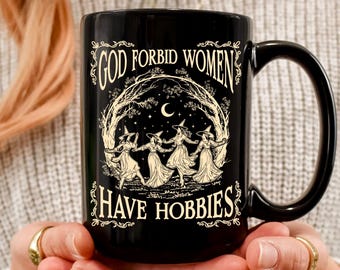 May include: A black ceramic mug featuring a cream-colored graphic of four figures in pointed hats dancing in a circle under a crescent moon and stars. The text "GOD FORBID WOMEN HAVE HOBBIES" is printed in a decorative font above and below the illustration.