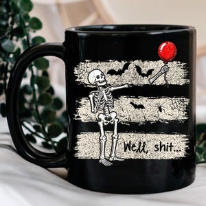 May include: A black ceramic mug featuring a graphic of a skeleton holding a red balloon. The design includes three horizontal distressed white stripes with bats, spiderwebs, and a spider. The text "Well, shit..." is printed in black.