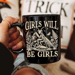 May include: A black ceramic mug featuring a cream-colored graphic of a praying mantis holding a heart, surrounded by botanical illustrations and small stars. The text "GIRLS WILL BE GIRLS" is printed in a serif font above and below the graphic.