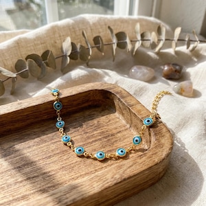 Könnte beinhalten: Ein goldfarbenes Kettenarmband mit mehreren kleinen, kreisförmigen Glücksbringer-Amuletten in Form von Augen. Jeder Anhänger besteht aus einer dunkelblauen Pupille, umgeben von einer hellblauen Iris und einem weißen äußeren Ring.