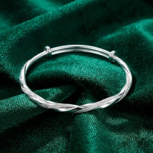 Può includere: Un bracciale rigido color argento dal design intrecciato, caratterizzato dall'alternanza di superfici lisce e texture scintillanti. Il bracciale circolare presenta due piccoli connettori metallici cilindrici sui lati opposti per una vestibilità regolabile.