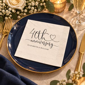 Pode incluir: Um guardanapo de papel quadrado branco com texto preto em cursivo que diz 40th anniversary, um pequeno ícone de coração e os nomes ELIZABETH & WILLIAM impressos abaixo. O guardanapo tem uma borda texturizada sobre um prato azul escuro.